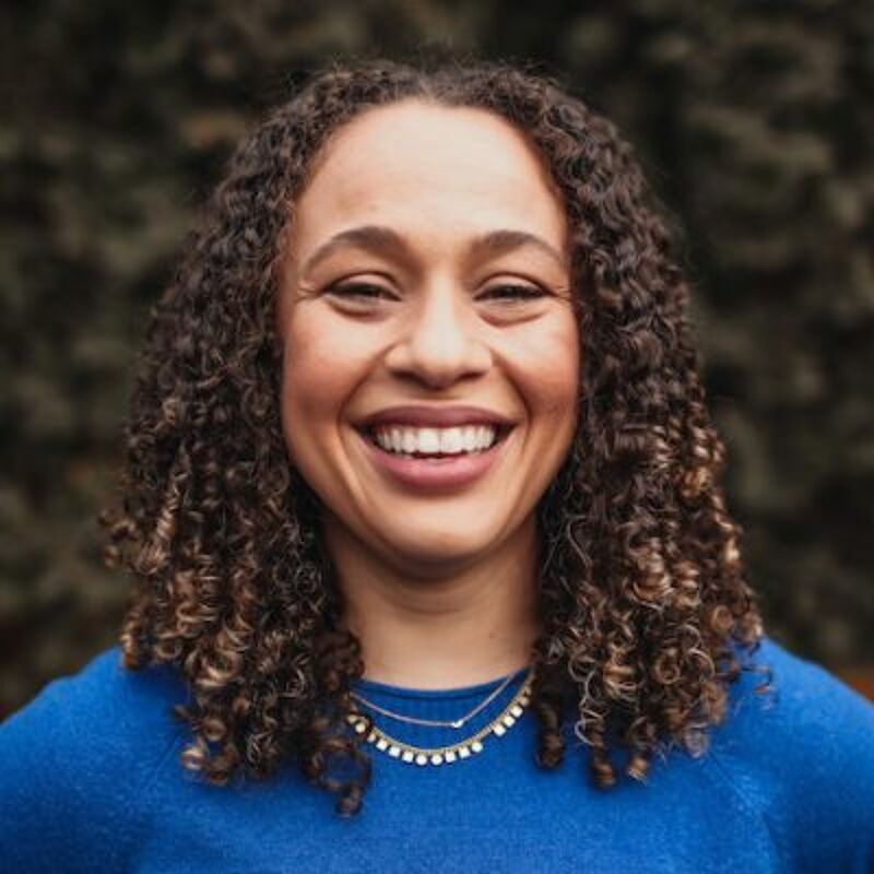 The image shows a woman with curly brown hair and a warm smile. She is wearing a blue sweater and a delicate necklace. The background is blurred, suggesting an outdoor setting, possibly a park or garden. Her expression is friendly and inviting.